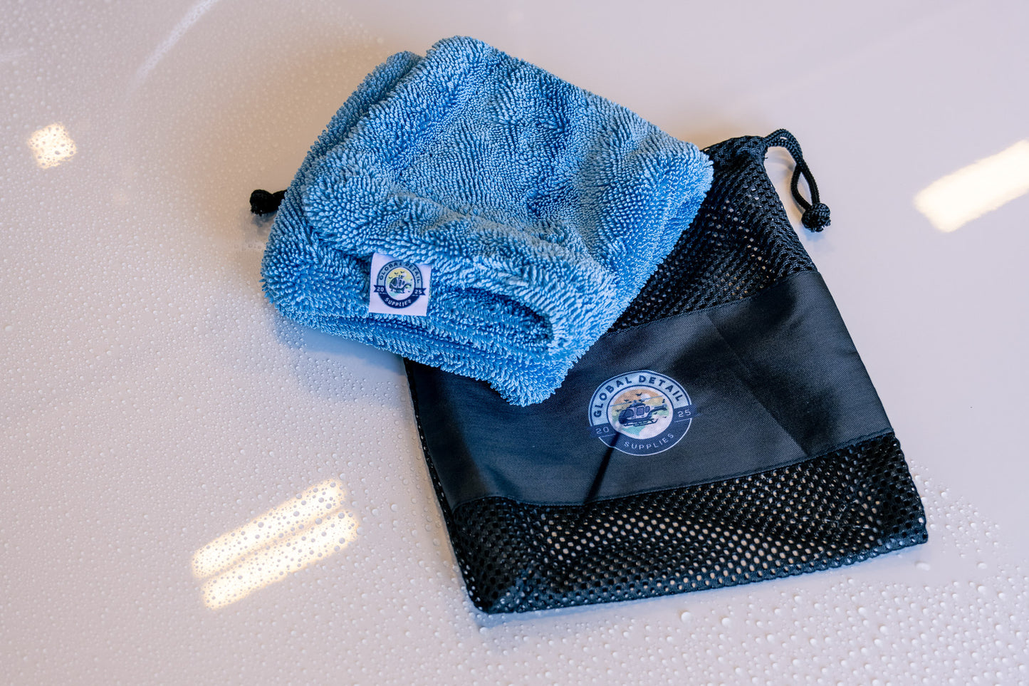 Blue microfiber AeroWipe Mini drying towel folded next to black mesh storage bag on a wet surface