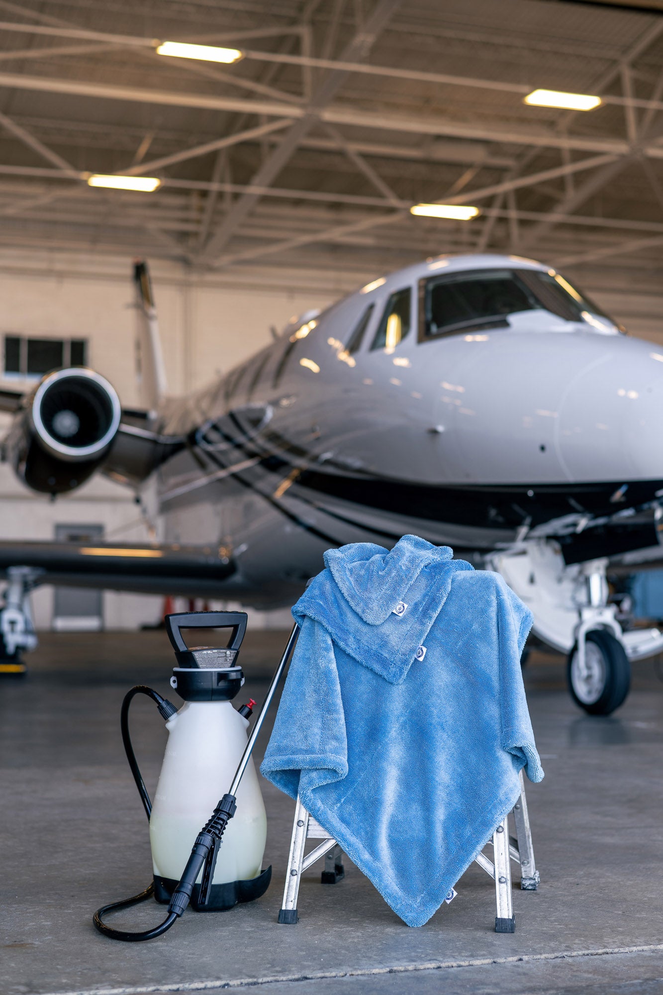 Blue microfiber towel draped over a small stool with a sprayer in front of a private jet inside an aircraft hangar