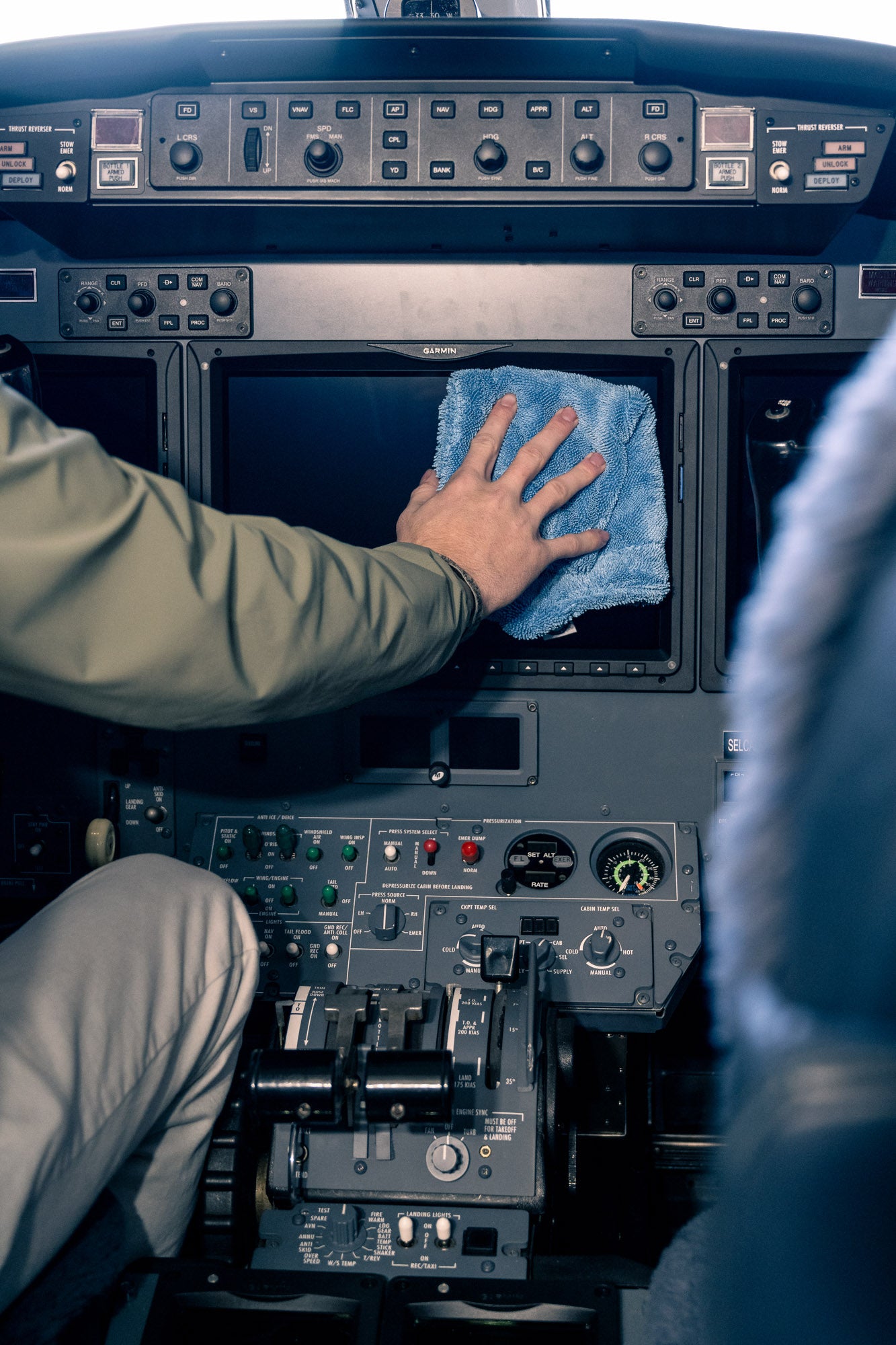 Person cleaning airplane cockpit screen with blue microfiber cloth wearing green jacket sleeve and beige pants