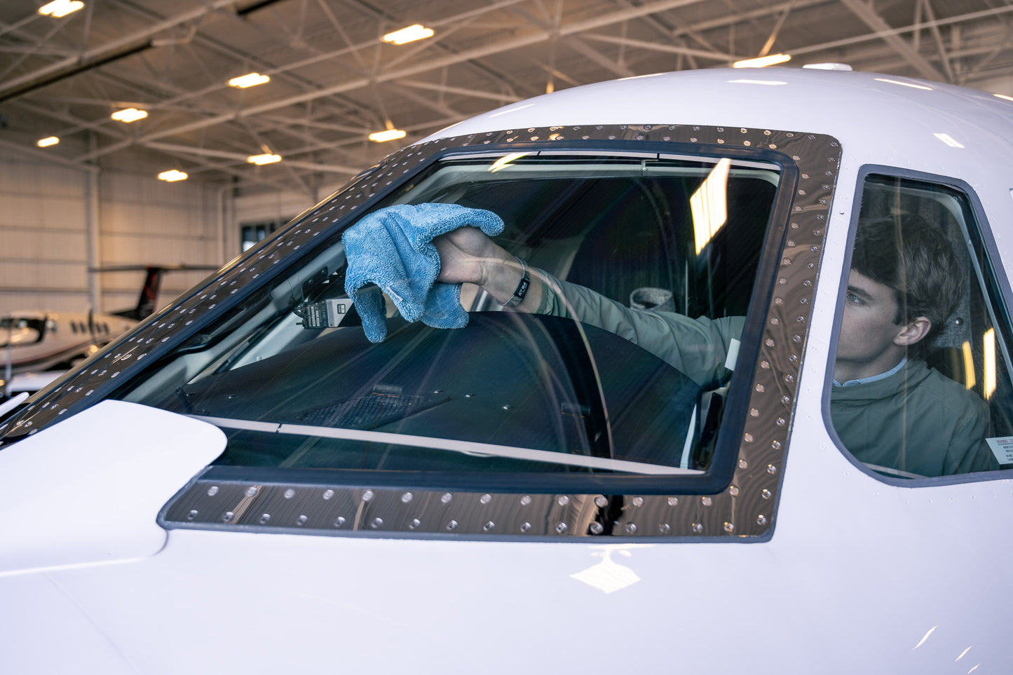Person using blue AeroWipe Mini microfiber towel to dry airplane cockpit window inside a hangar environment