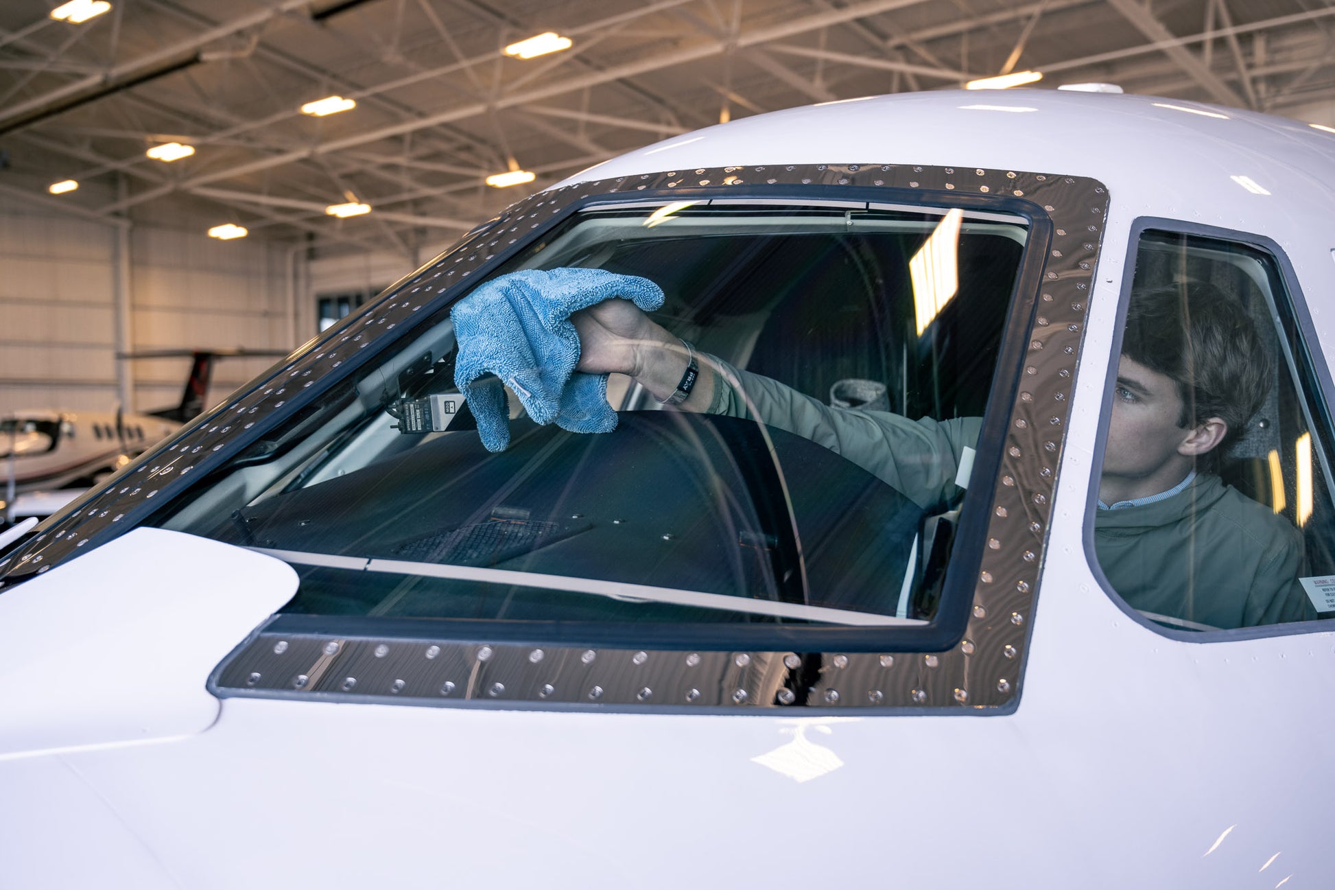Person using blue AeroWipe Mini microfiber towel to dry airplane cockpit window inside a hangar environment