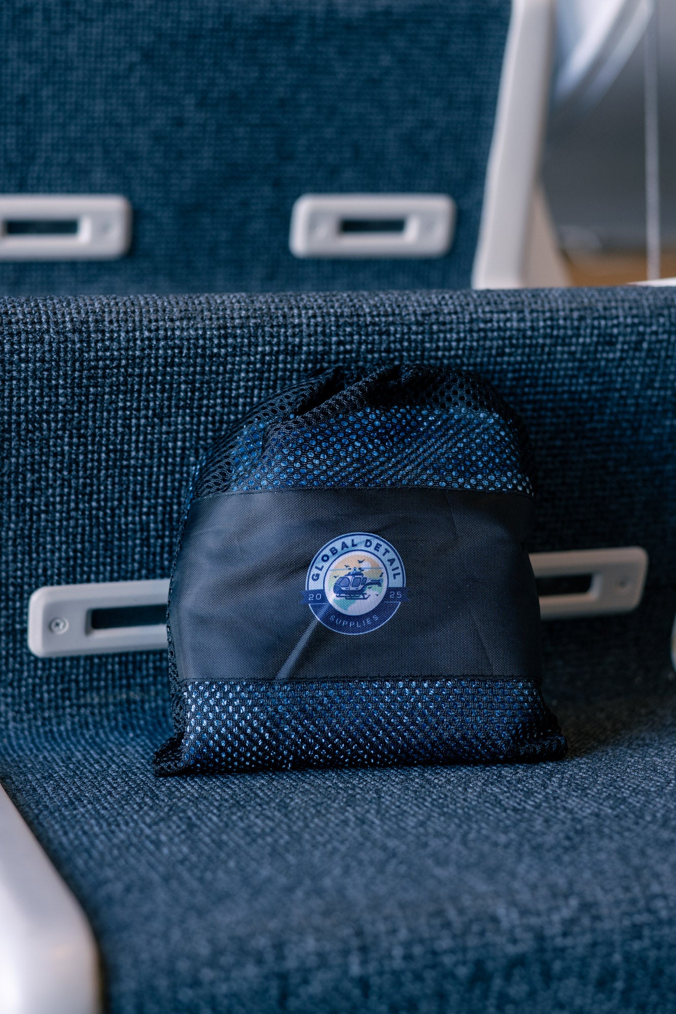 Black mesh drawstring bag with Global Detail Supplies logo sitting on dark blue textured airplane seat